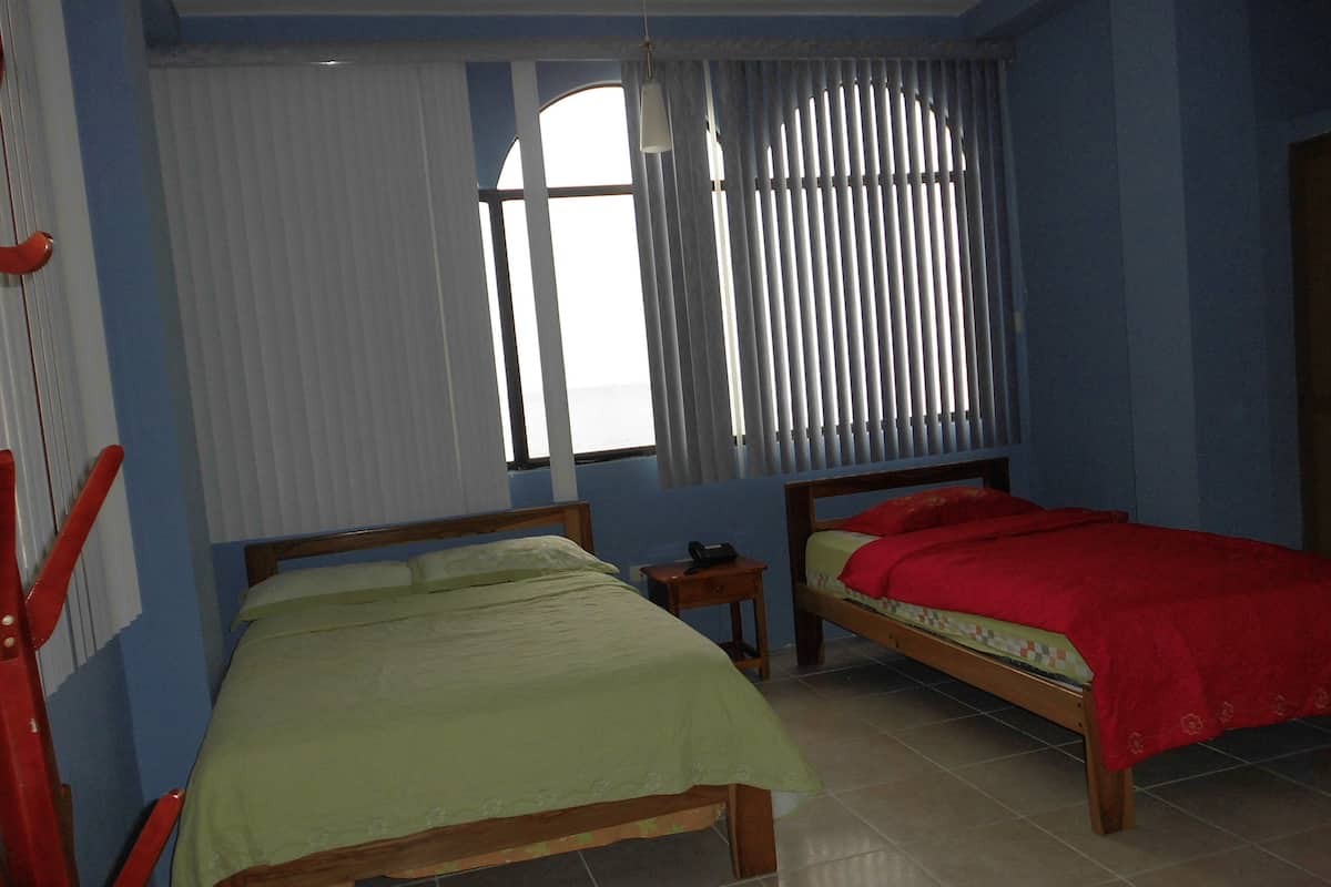 Una habitación con dos camas de madera, una ventana con persianas, una silla roja y una mesita con un teléfono.