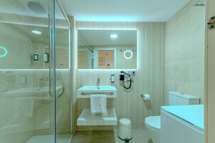 Une salle de bain moderne avec une douche à l’italienne en verre, un lavabo blanc avec une serviette, des toilettes et un miroir doté d’un anneau lumineux.