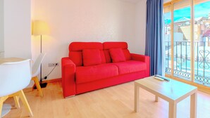 A red sofa, a white chair, a small table, a lamp, and a view of a balcony with metal railings. - Apartamentos Globus by Be Alicante (Alicante)