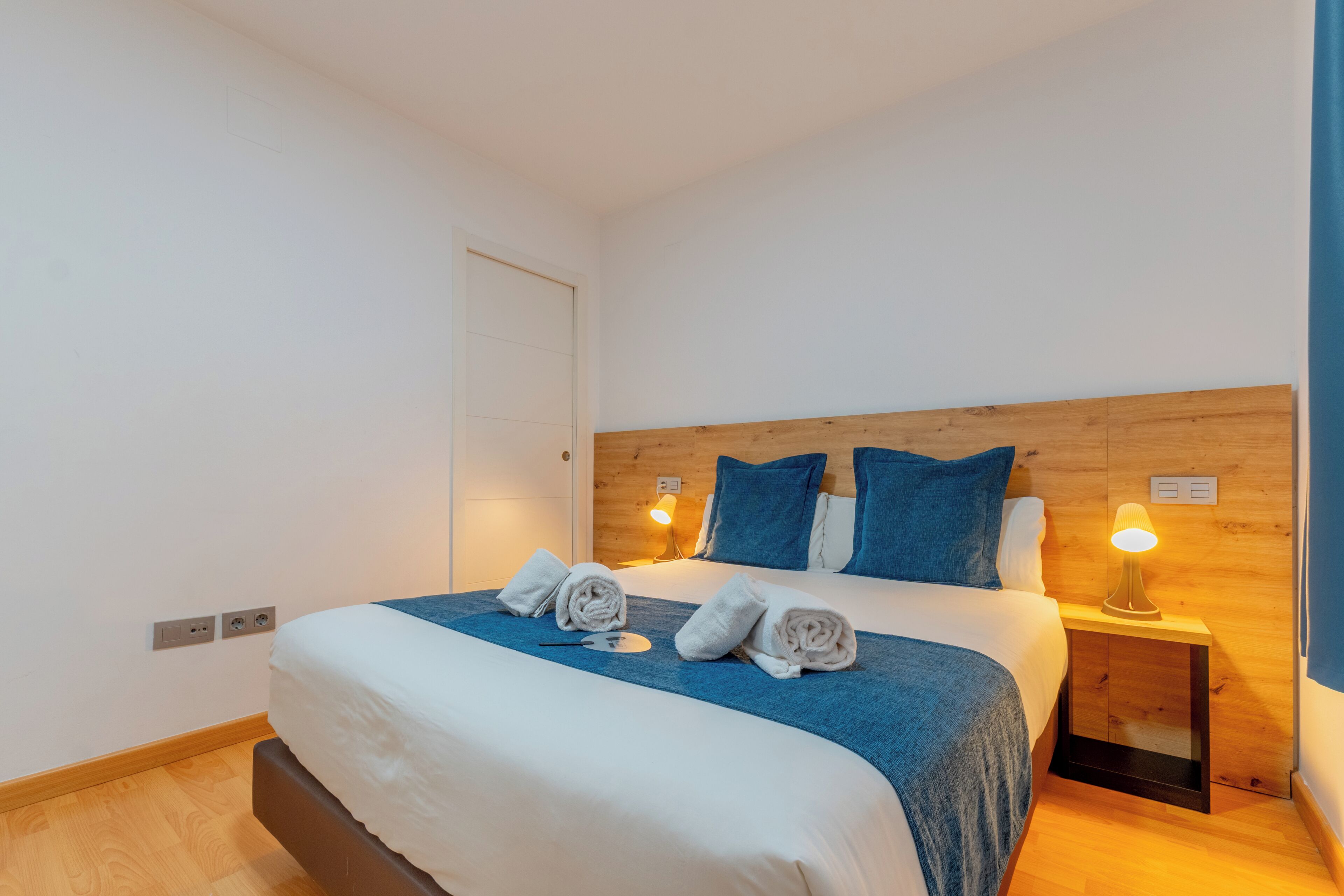 Una cama bien hecha con una manta azul, dos almohadas y dos toallas enrolladas. Hay dos lámparas de noche sobre mesitas de madera.
