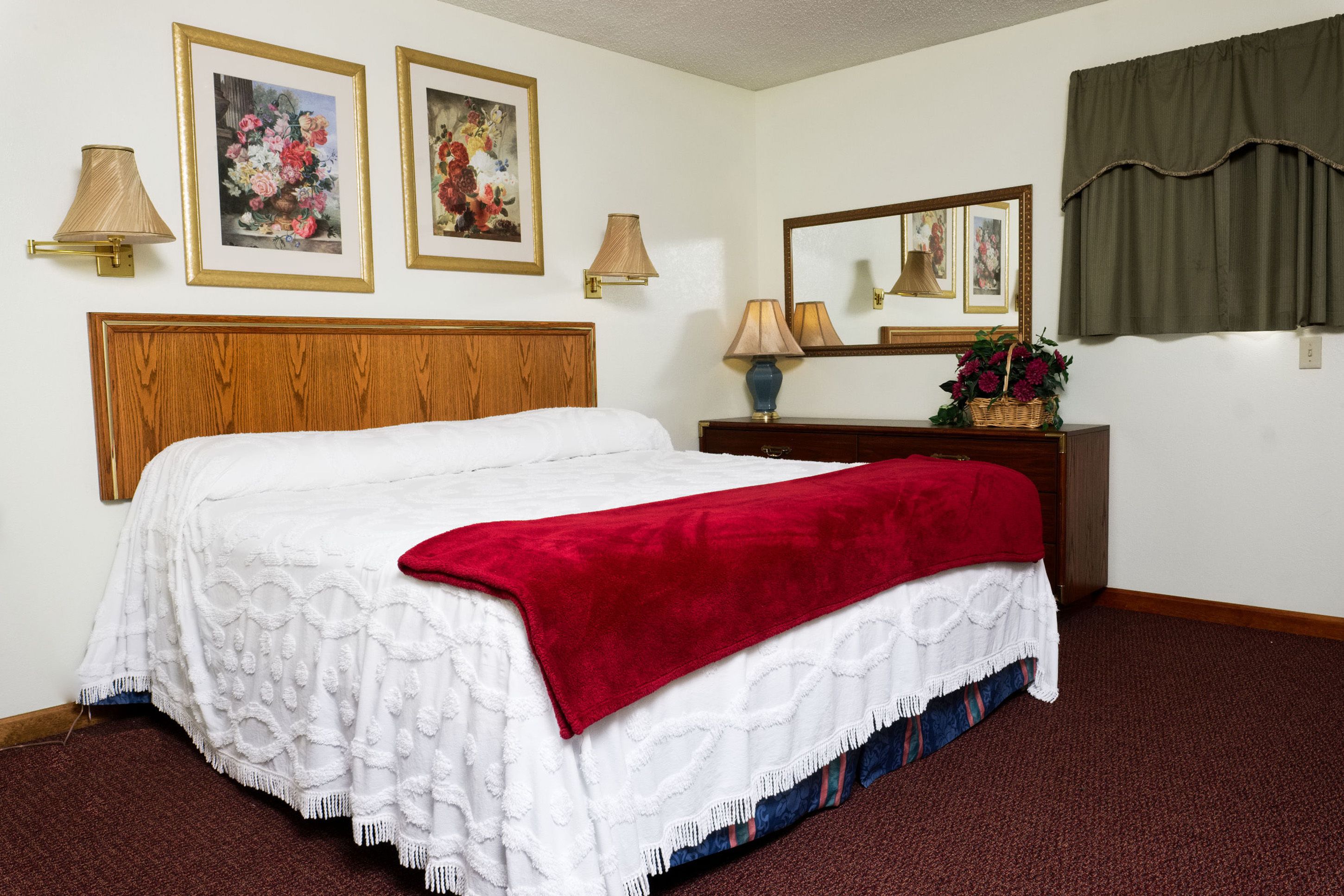Una habitación de hotel con cabecera de madera, cama blanca con una manta roja, cómoda de madera, espejo y dos cuadros florales enmarcados en la pared.
