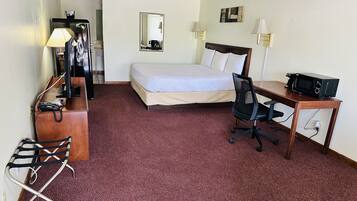 A hotel room with a bed, desk, chair, desk lamp, and a microwave.