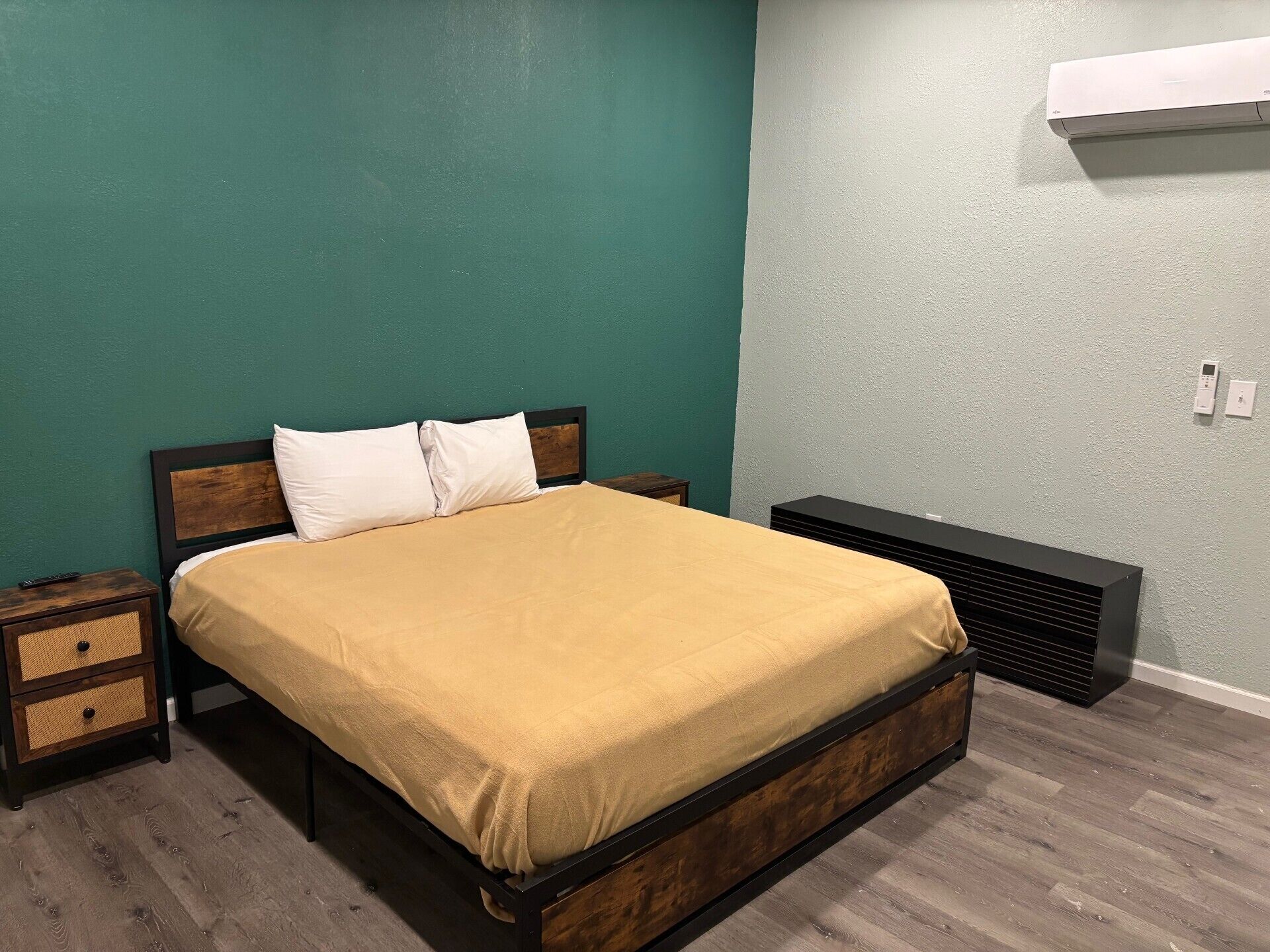 A bed with a wooden frame and a beige bedspread, a bedside table with a woven basket, and a wall-mounted air conditioner.