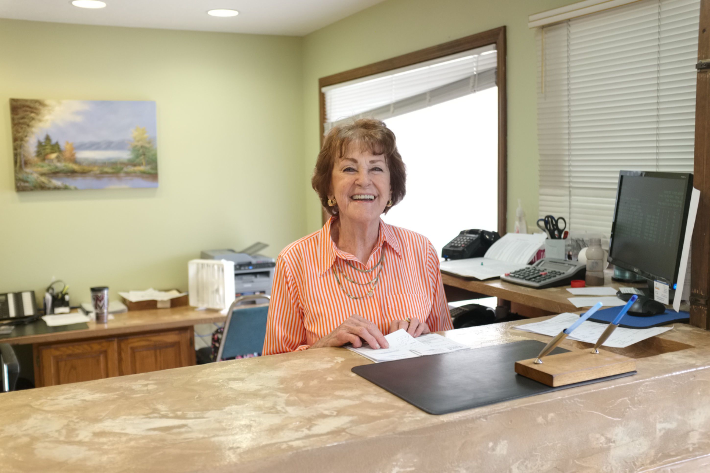 A woman seated at a desk in an office with a computer, telephone, and various office supplies.