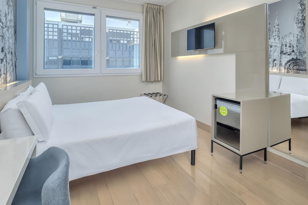 A hotel room with a bed, a desk, a chair, a TV, and a bathroom mirror.