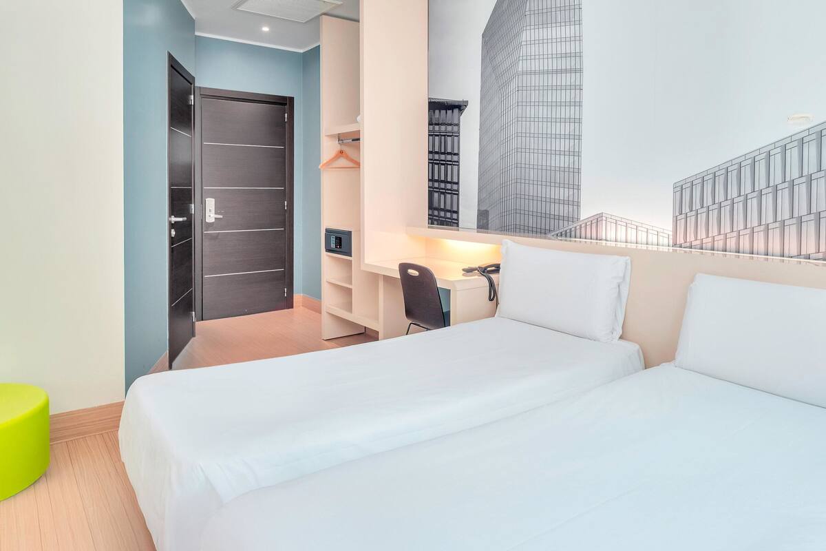 A hotel room with a bed, a desk, a chair, a wardrobe, and a view of a modern building.