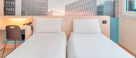 A hotel room with two single beds, a desk with a chair, and a view of modern buildings.