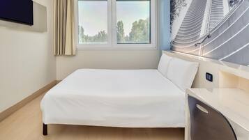 Una habitación de hotel con cama, televisión fijada a la pared, ventana con cortinas y lámpara de pared.