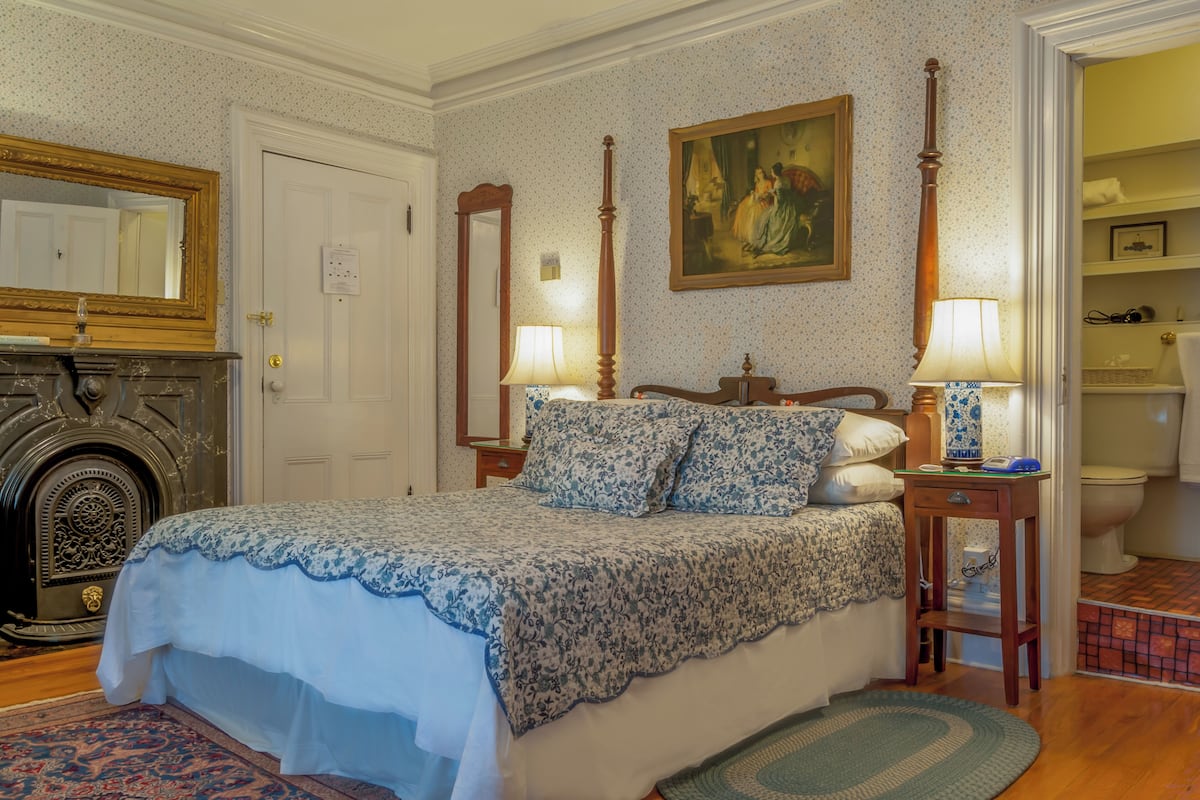A bedroom with a bed, two bedside tables with lamps, a painting on the wall, a door, and a fireplace.