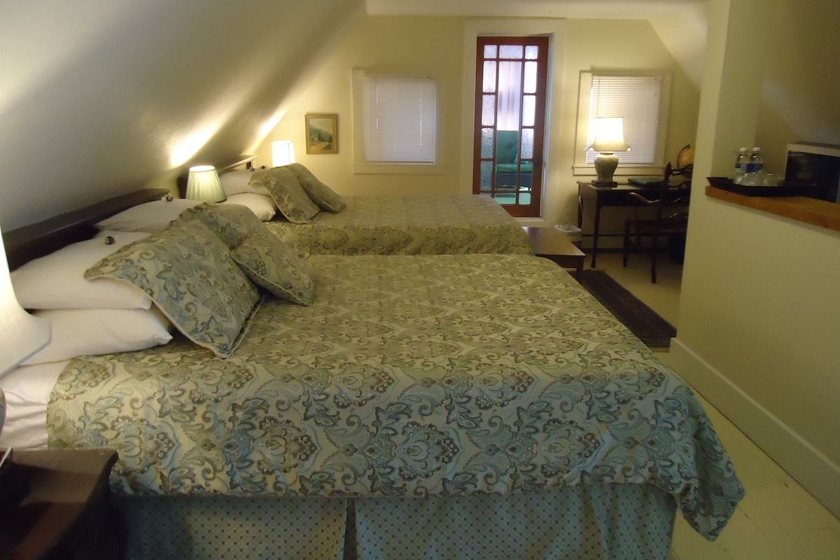 A bedroom with two beds, a desk, and a chair. There are two windows with blinds, a door, and a picture on the wall.