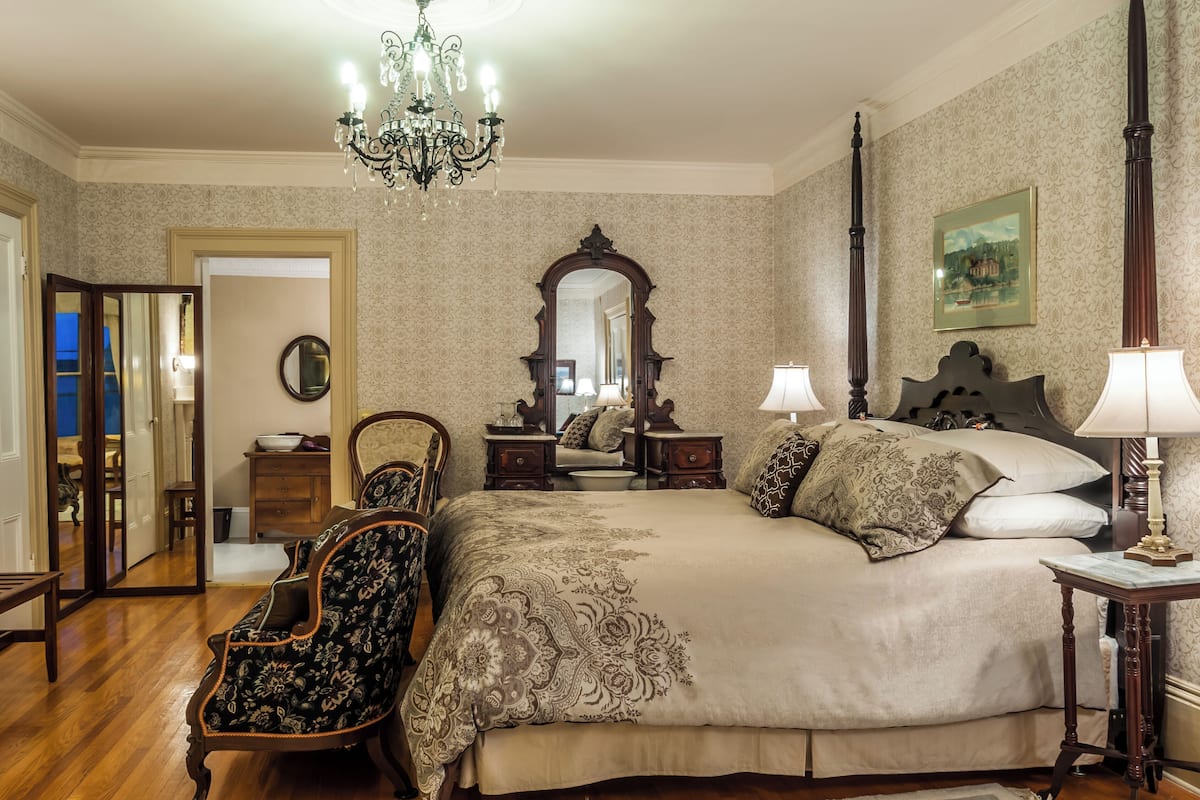 A bedroom with a large bed, two bedside tables, a chandelier, a mirror, and a chair.