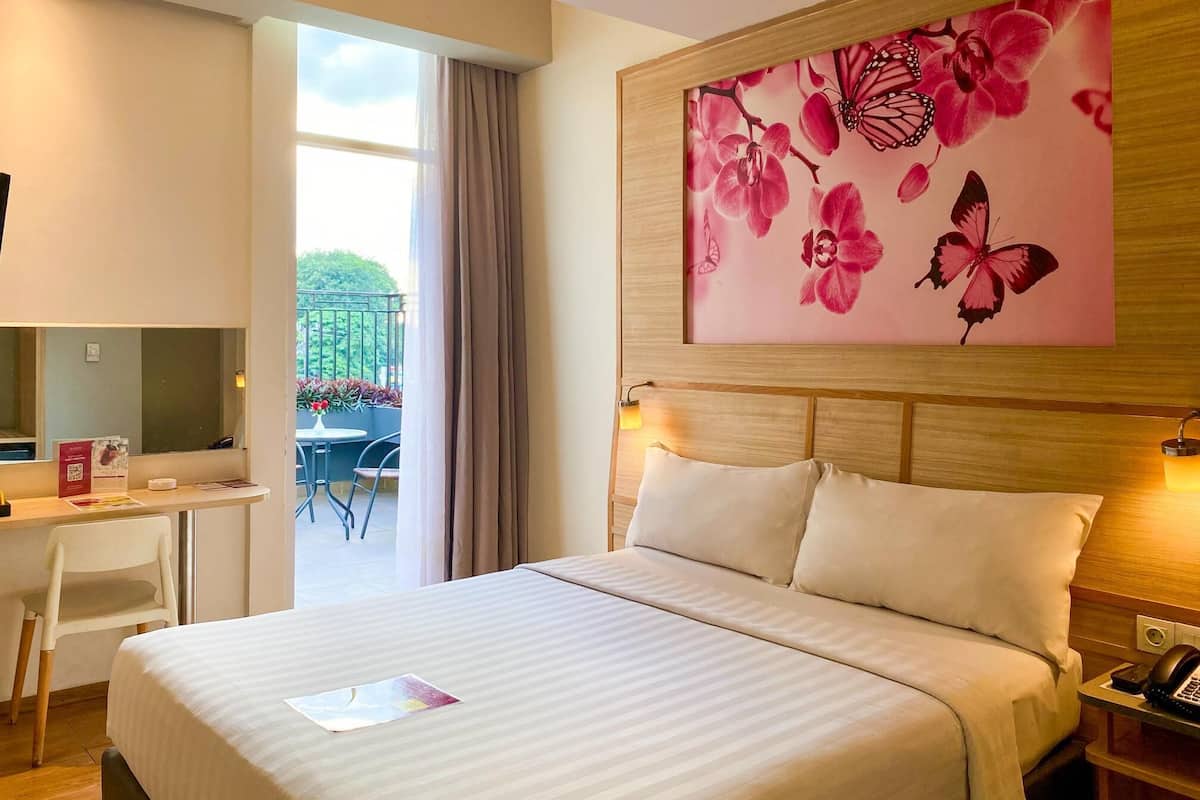 A hotel room with a bed, a desk, a chair, a bedside table with a phone, and a large floral artwork on the wall.