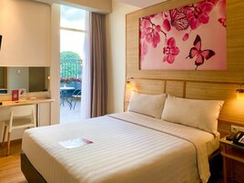 A hotel room with a bed, a desk, a chair, a bedside table with a phone, and a large floral artwork on the wall.