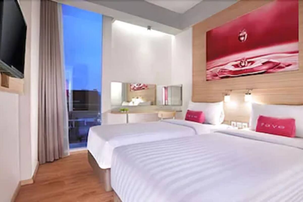 A hotel room with two beds, a TV, a desk, and a large window.