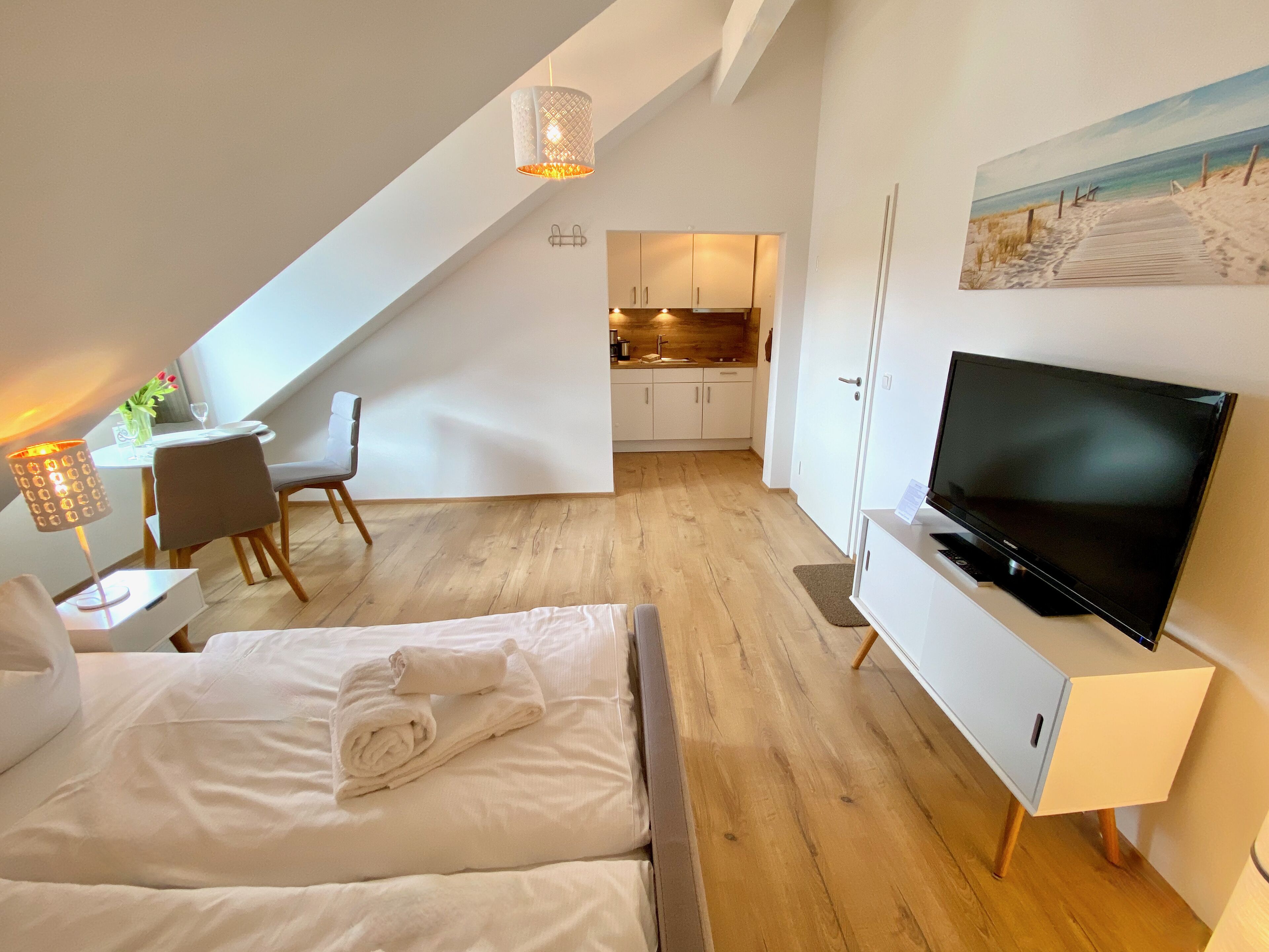 A hotel room with a bed, a TV, a dining area, and a kitchen in the background.