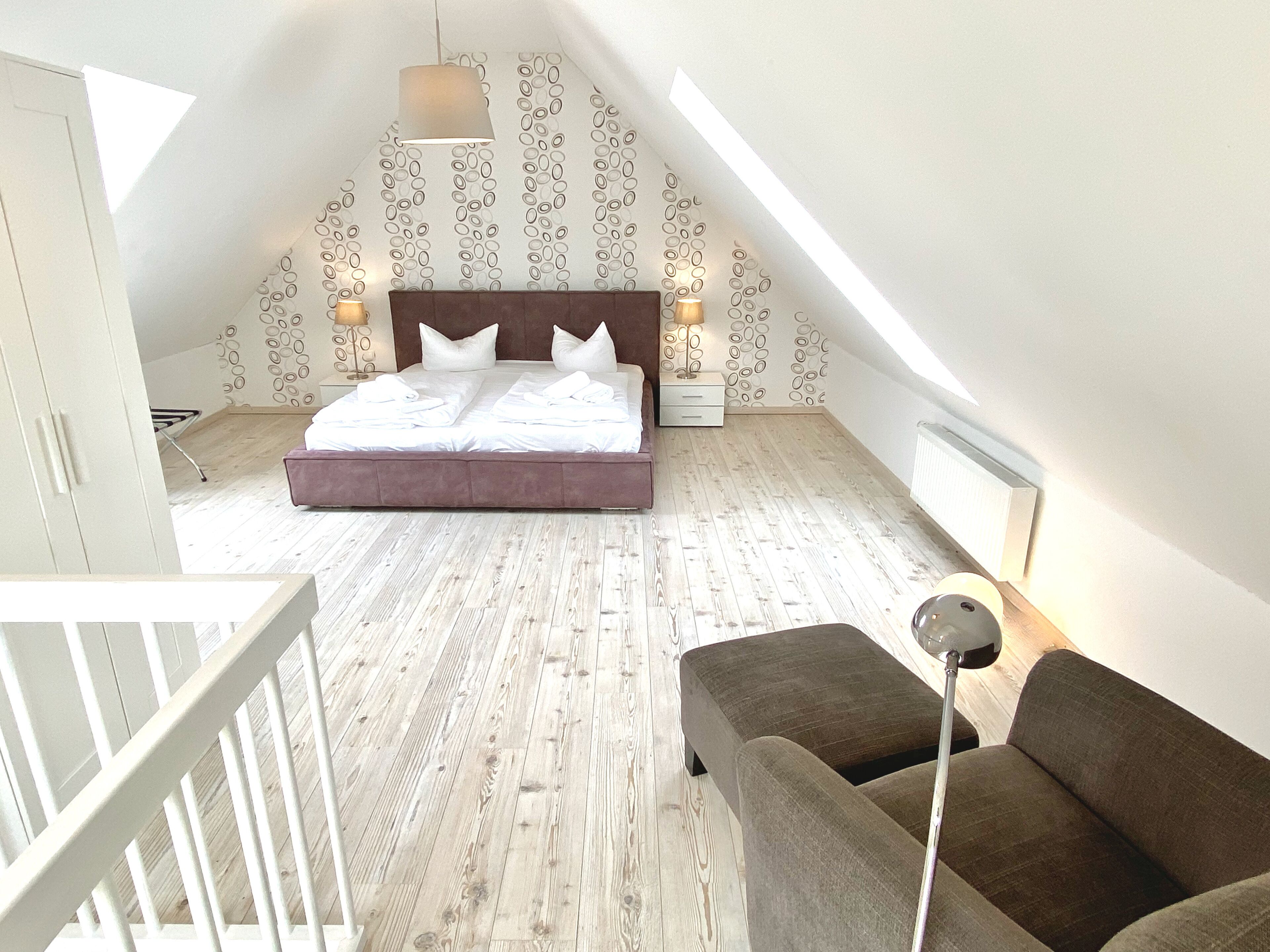 A bedroom with a large bed, a nightstand, a lamp, a chair, and a wooden floor.