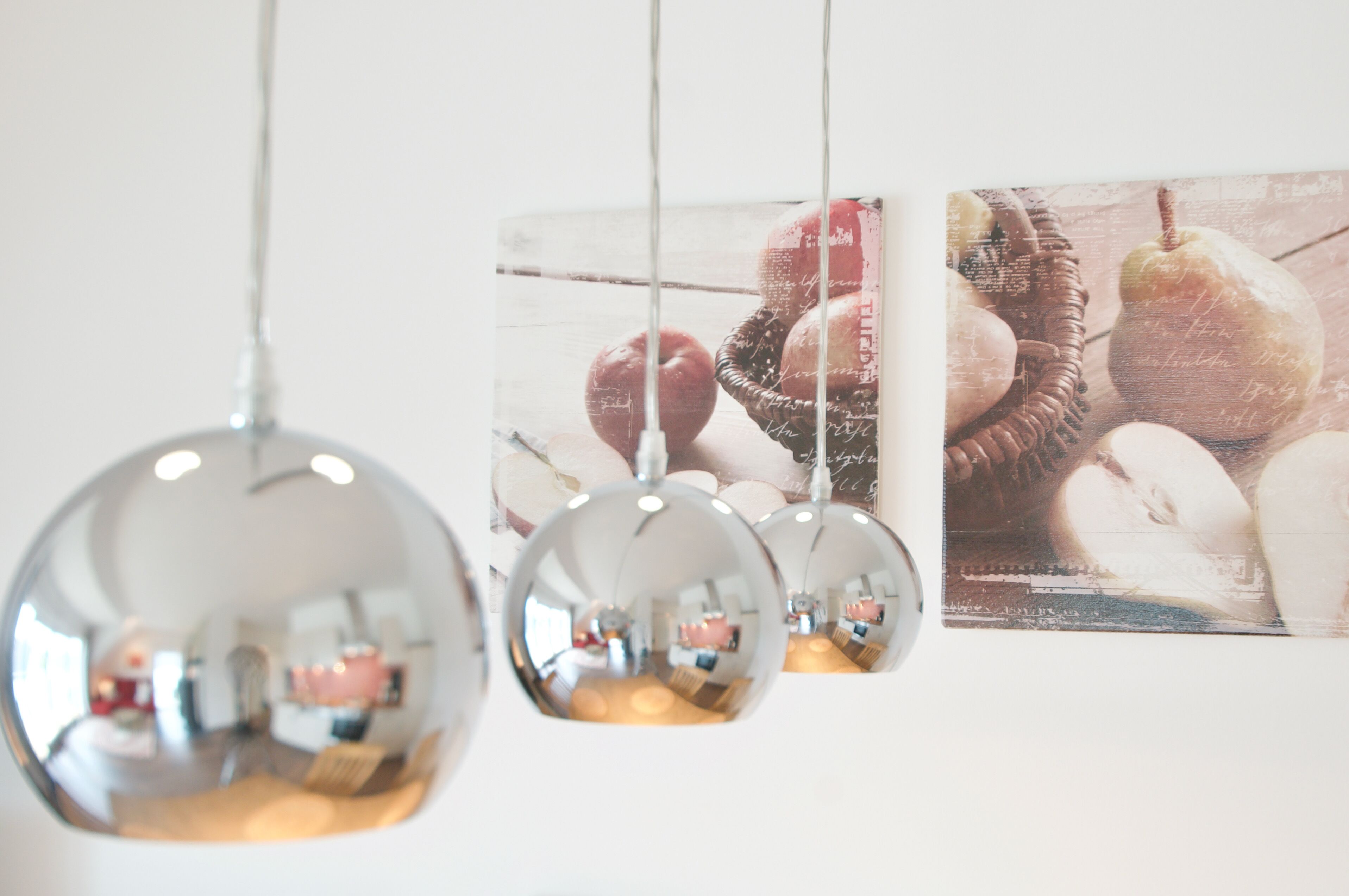 Una habitación con un espejo grande que refleja una zona de comedor, un cuadro colgado con frutas y un objeto esférico colgante.