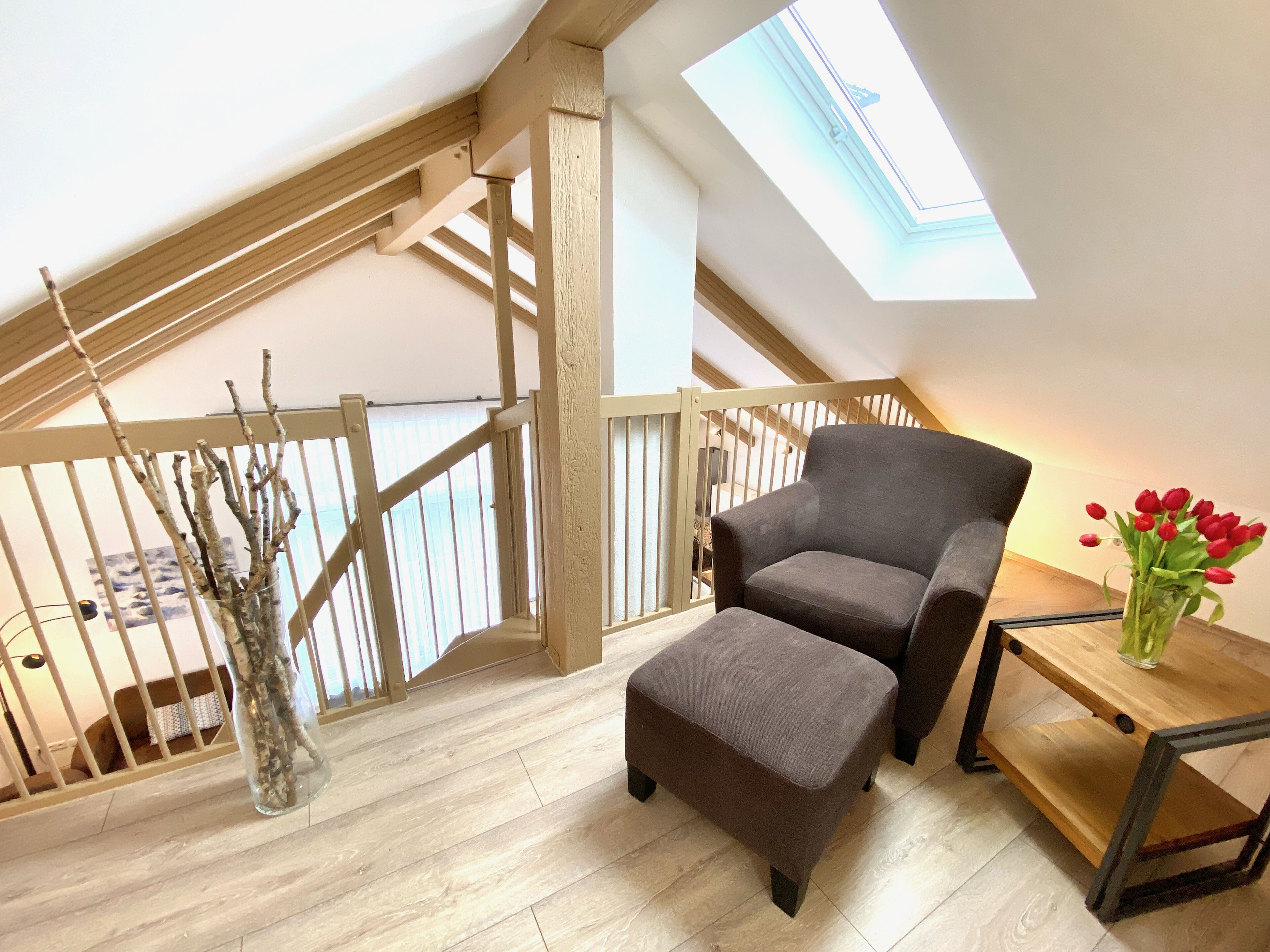 Ein Loft-Zimmer mit hoher Decke, Holzbalken, einem Oberlicht, einem grauen Sessel, einem kleinen Hocker, einem Holztisch mit einer Vase roter Blumen und einer Glasvase mit Zweigen.