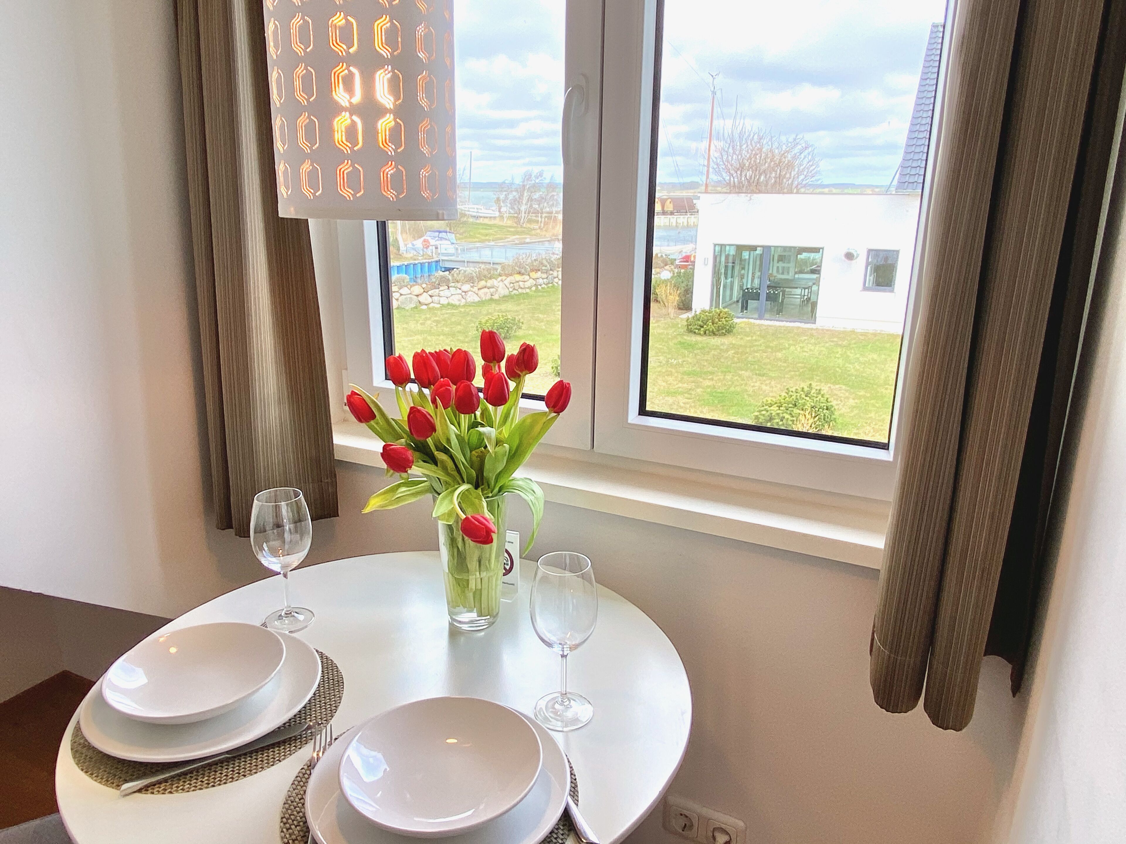 Une table à manger dressée avec de l’assiette blanche, des couverts et un vase rempli de tulipes rouges.