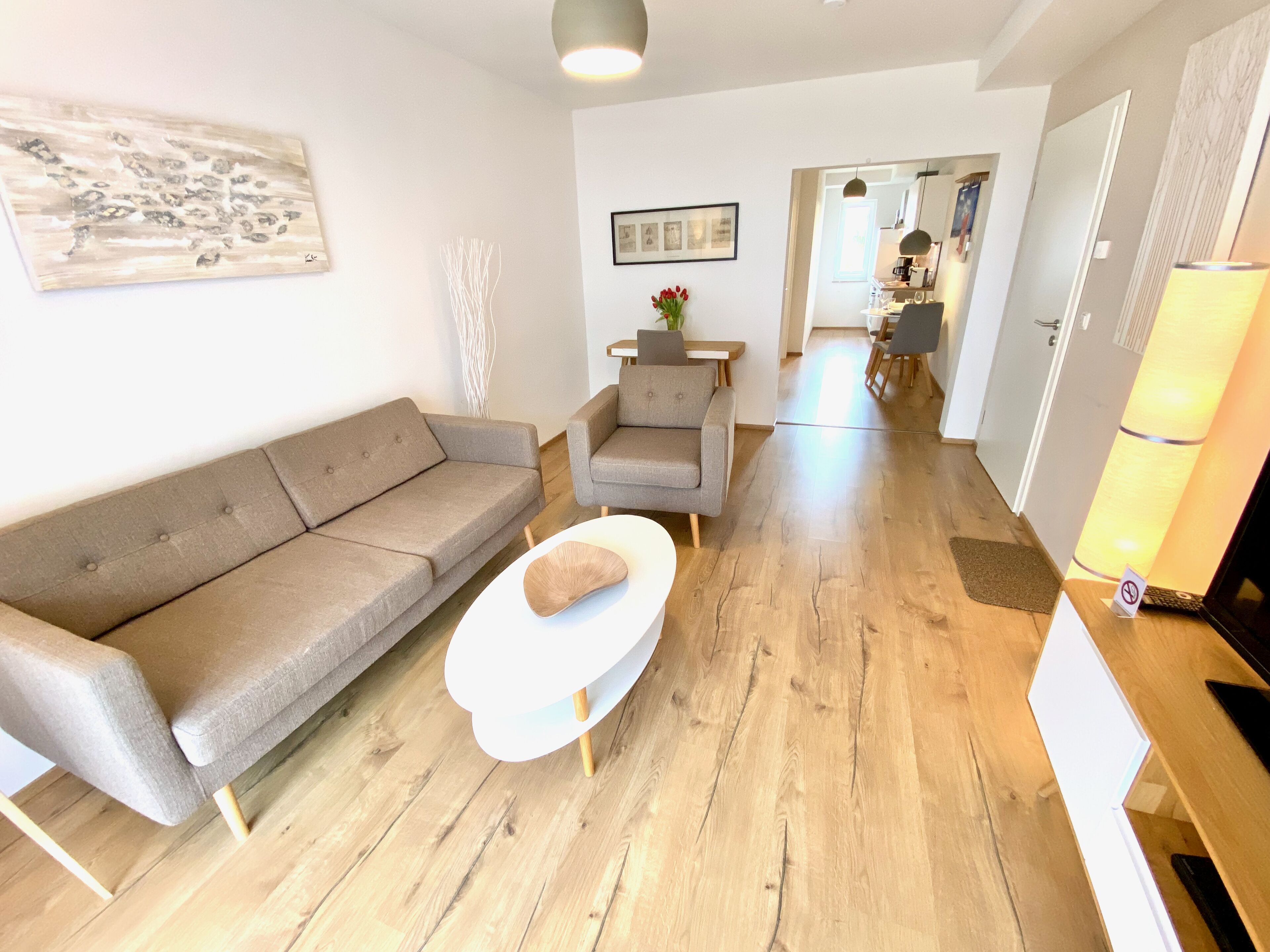 Un salon moderne avec un canapé gris, une table basse blanche et un plancher en bois. Un tableau encadré est accroché au mur et une lampe se trouve sur la droite.