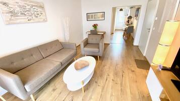 Un salon moderne avec un canapé gris, une table basse blanche et un plancher en bois. Un tableau encadré est accroché au mur et une lampe se trouve sur la droite.