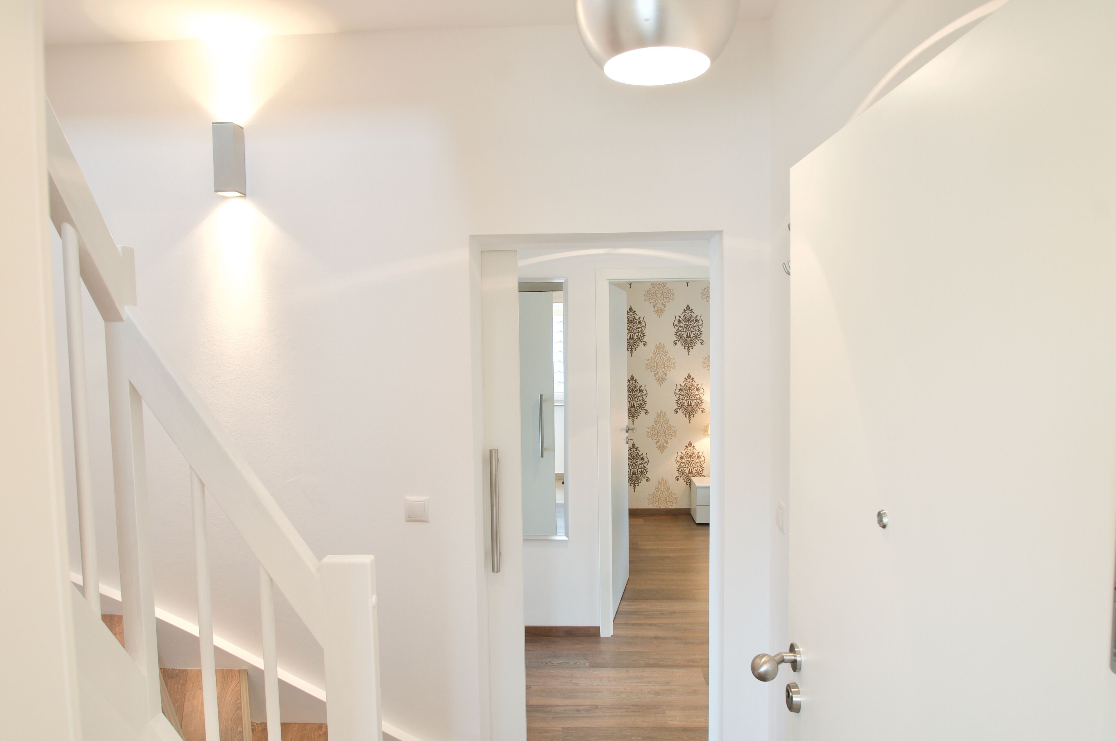 A hallway with white walls, a wooden floor, and a door leading to a room with patterned wallpaper.