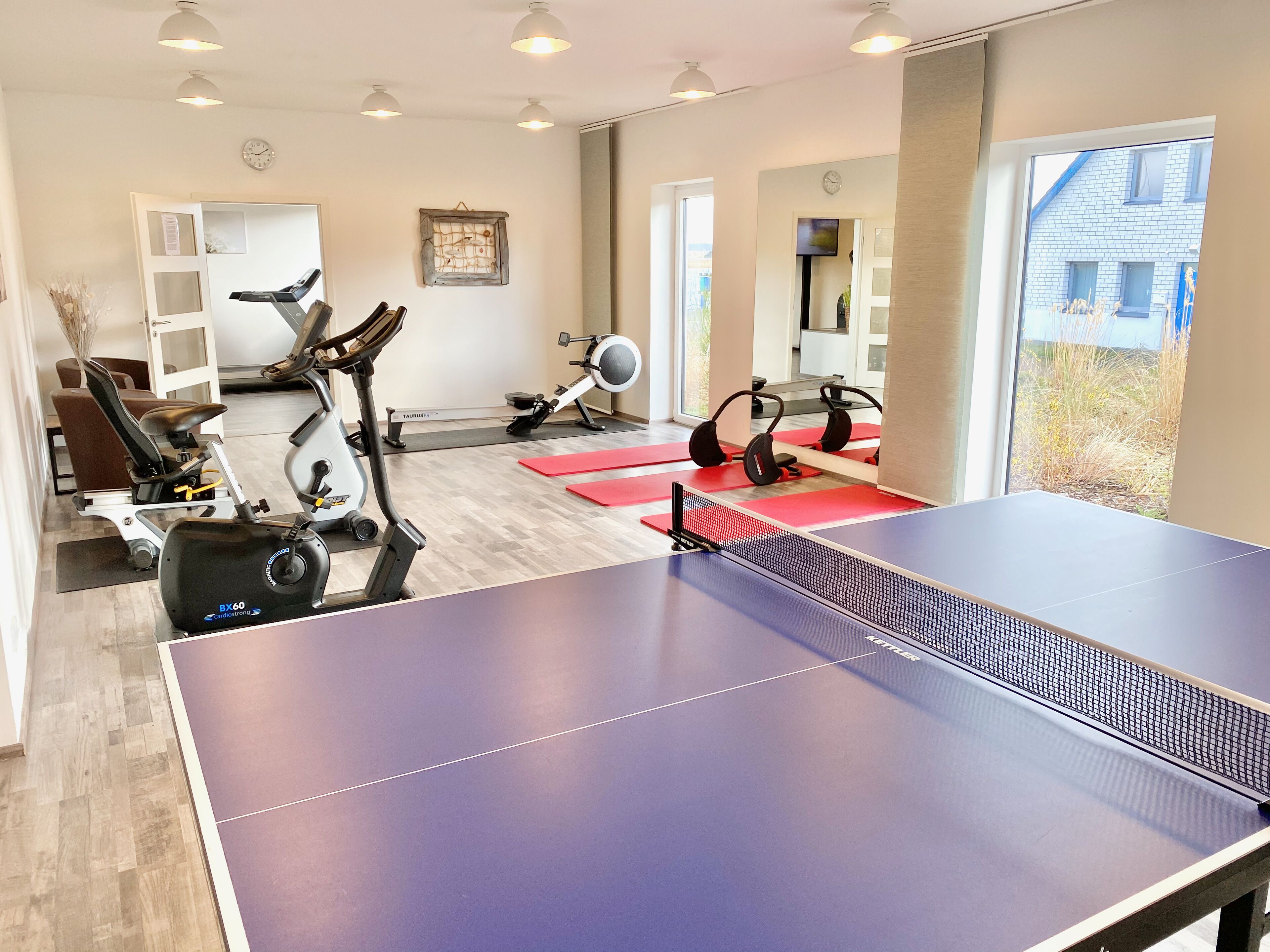 Ein Fitnessraum mit Trainingsgeräten, einem Tischtennis-Tisch und großen Fenstern.