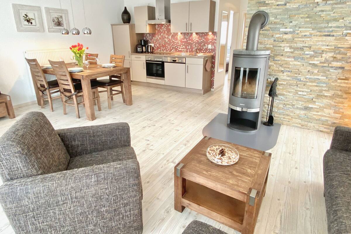 A modern living room with a fireplace, a dining area with wooden furniture, and a kitchen with a stone backsplash.