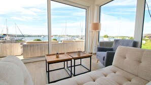A modern living room with a large window overlooking a marina, featuring a sofa, a coffee table, and a lamp. - Moenchgut Living & Spa (Mönchgut-Granitz)