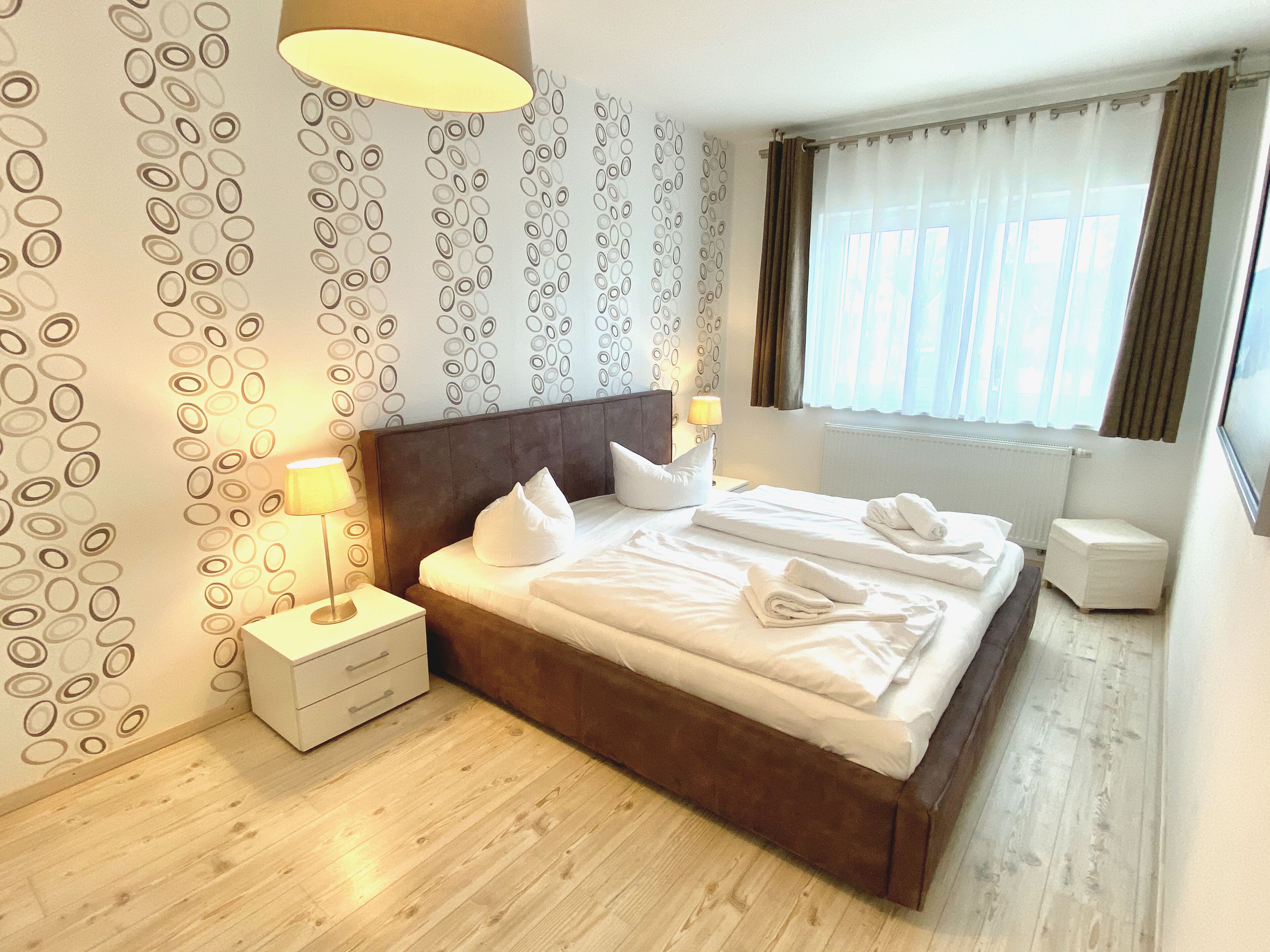 Een net opgemaakt bed met kussens en handdoeken, een bedlampje, een nachtkastje en een raam met gordijnen.