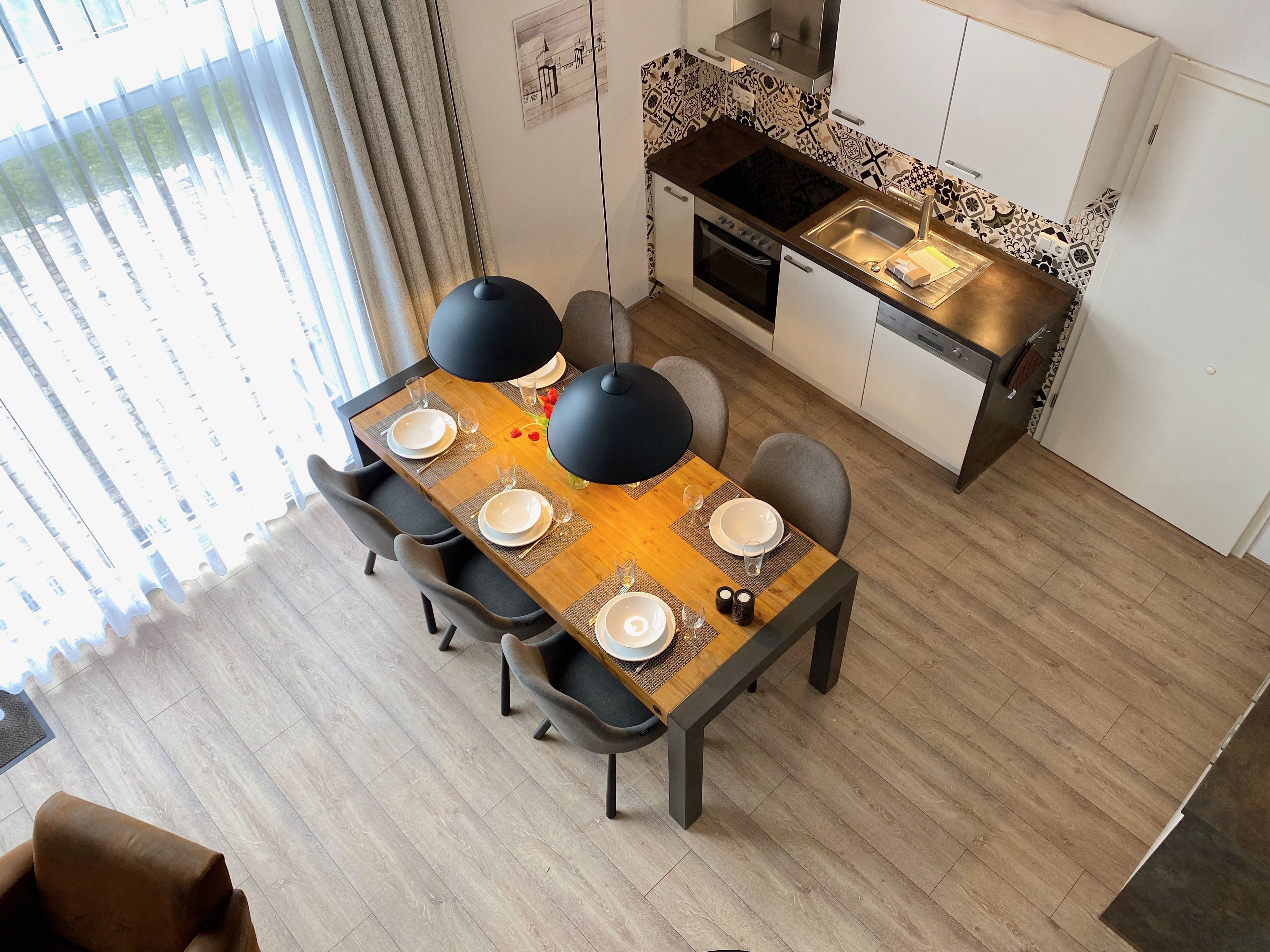 Un comedor con mesa de madera dispuesta para cuatro, una cocina con salpicadero decorado y un ventanal con cortinas.