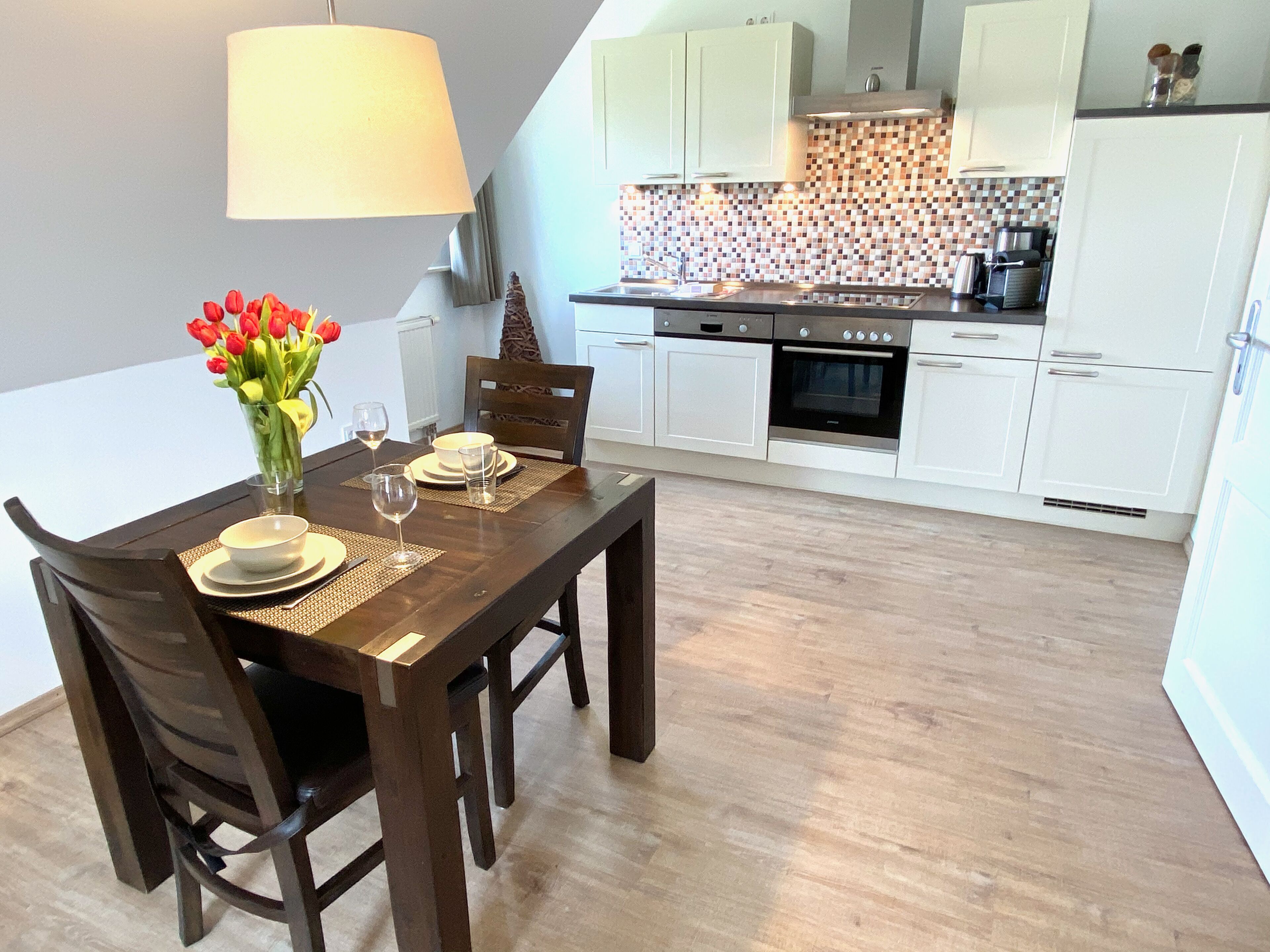 Een eethoek met een houten tafel gedekt voor vier personen, een vaas rode tulpen en een lamp.