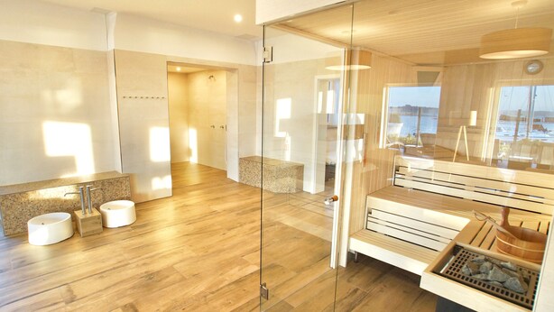 A modern bathroom with a glass shower enclosure, wooden flooring, and a view of the sea through the window. - Moenchgut Living & Spa (Mönchgut-Granitz)