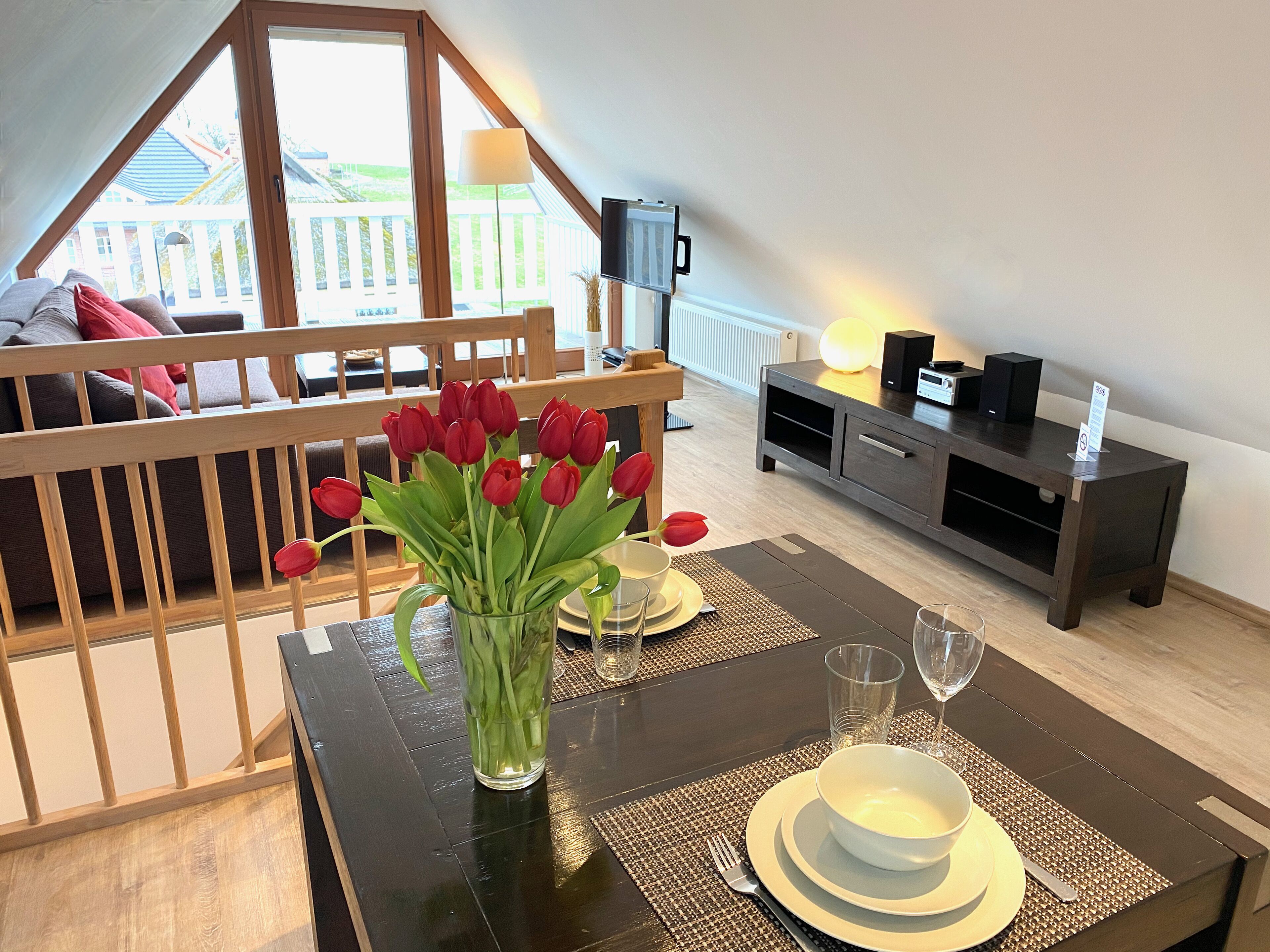 A dining area with a table set for two, a vase of red tulips, and a view of a wooden deck and white fence.