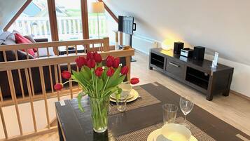 A dining area with a table set for two, a vase of red tulips, and a view of a wooden deck and white fence.