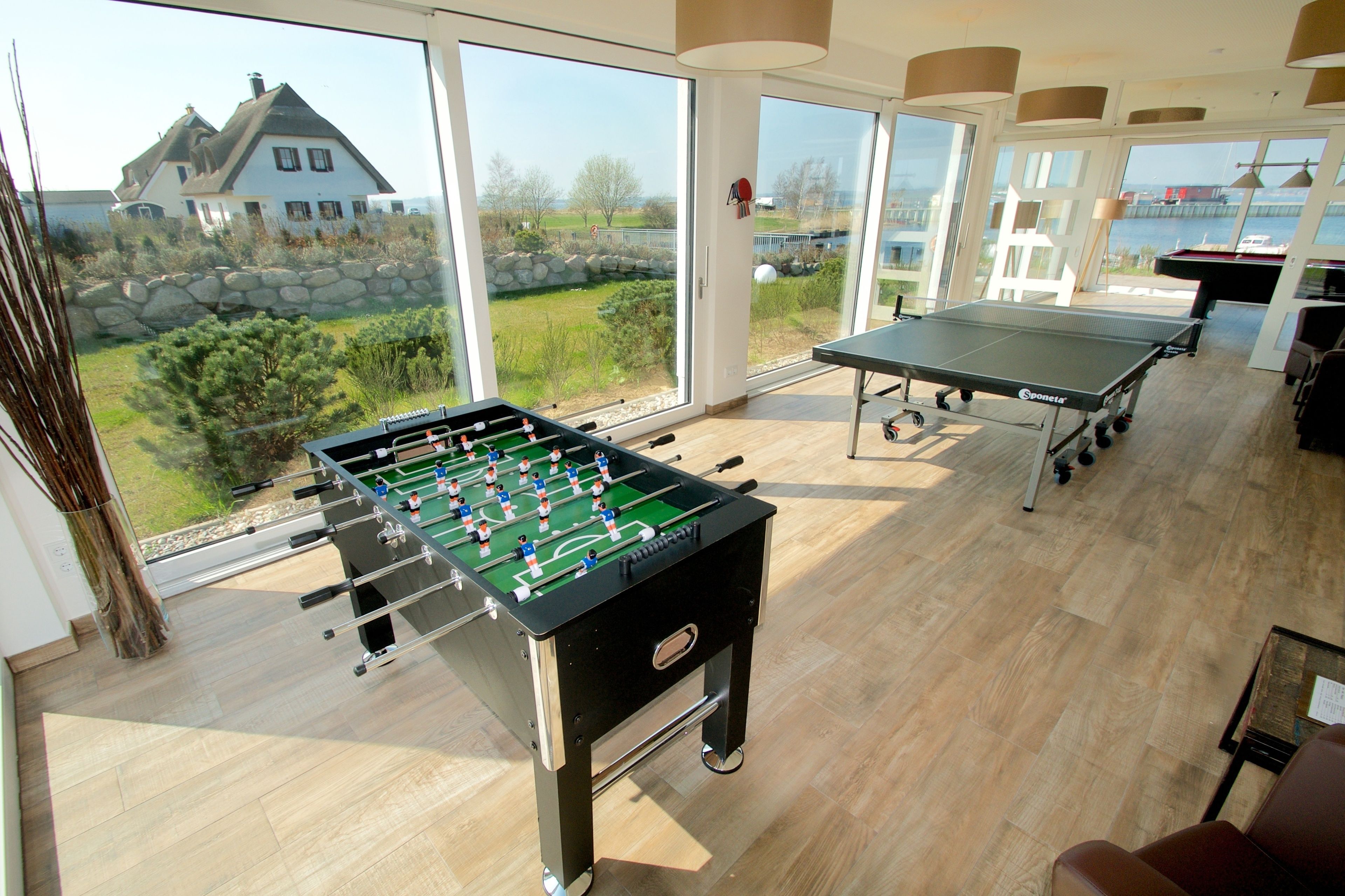 Una habitación con mesa de fútbol de mesa, mesa de ping pong y vistas a una casa y paisaje a través de amplios ventanales.