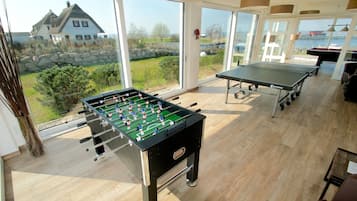 Una habitación con mesa de fútbol de mesa, mesa de ping pong y vistas a una casa y paisaje a través de amplios ventanales.