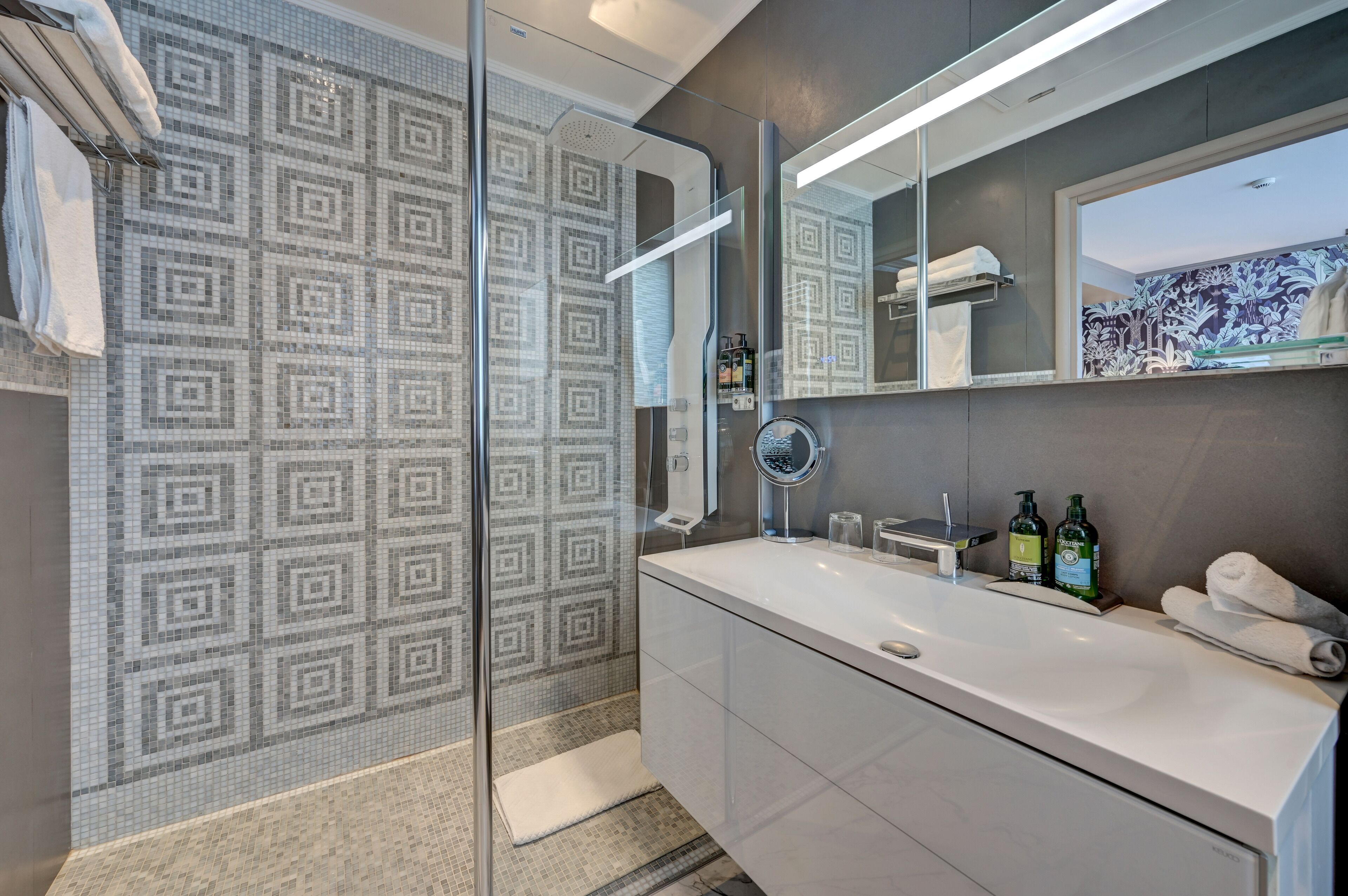 A modern bathroom with a patterned shower wall, a large mirror, and a white vanity with toiletries.