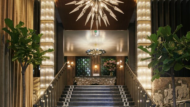 A grand staircase with a modern chandelier, a stone wall with a floral arrangement, and a large vase at the bottom. - The One Alma Paris (Paris)