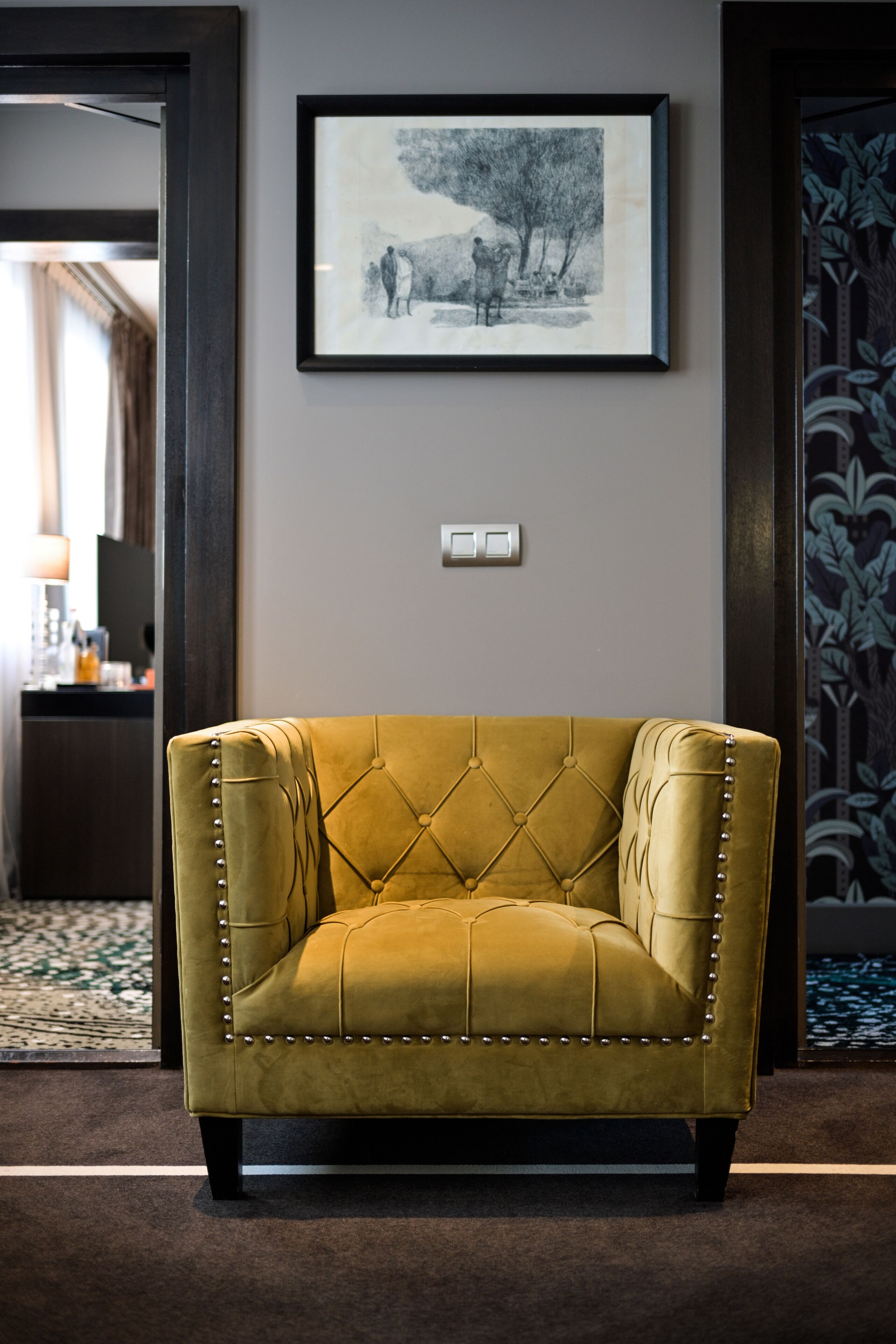 A mustard-colored tufted armchair with metal studs, positioned in a hallway with a framed black-and-white artwork above it.