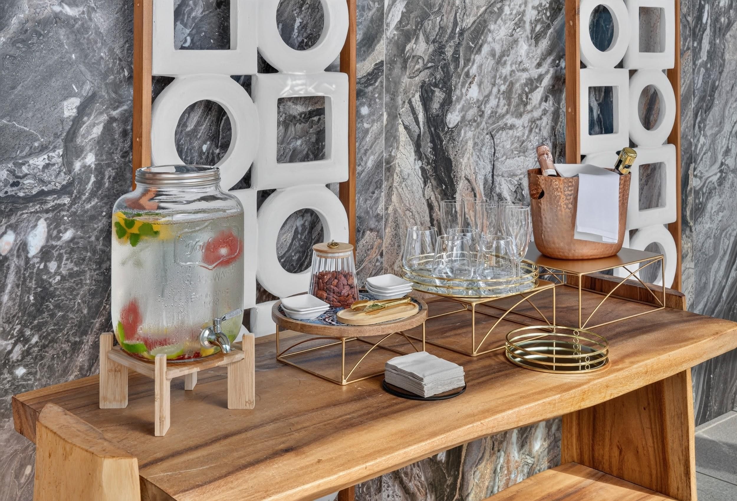 A wooden table with a glass jar of iced drink, a copper mug, and a decorative metal tray with glasses and plates.