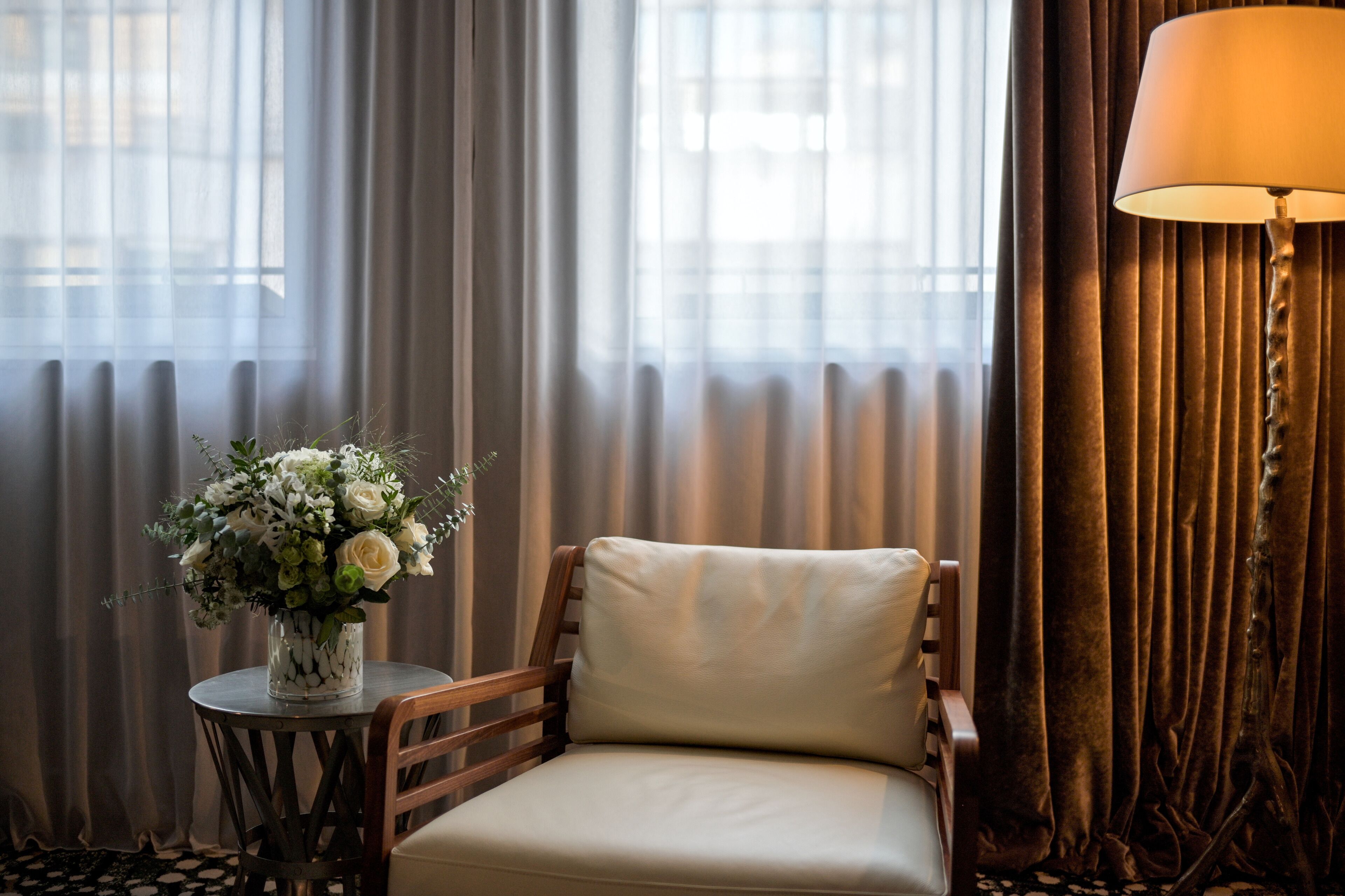 A room with a chair, a small table with a vase of flowers, and a lamp.