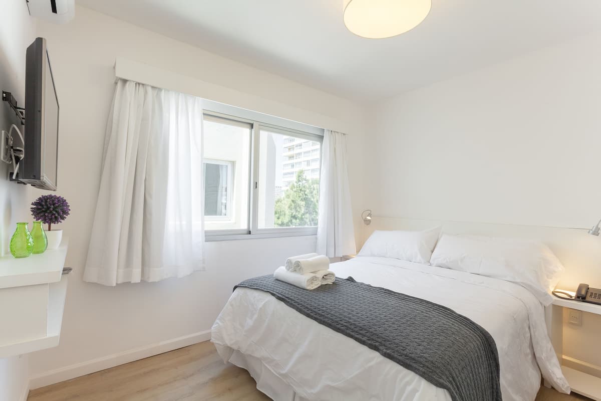 Un dormitorio con cama, mesita de noche, lámpara, ventana con cortinas y televisor montado en la pared.