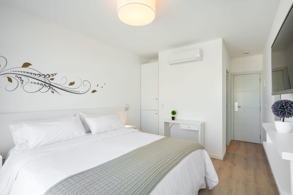 Un dormitorio moderno con una cama grande, una mesita de noche con una planta, un televisor montado en la pared y una puerta que da a otra habitación.