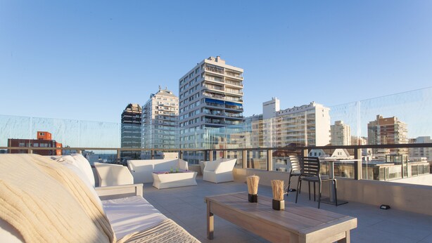 Una terraza en la azotea con un sofá, una mesa de madera y sillas. Se ven edificios al fondo.