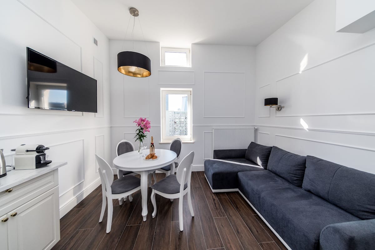 Una sala de estar moderna con un sofá gris, una mesa de comedor con sillas, un televisor de pantalla plana montado en la pared y una lámpara colgante.