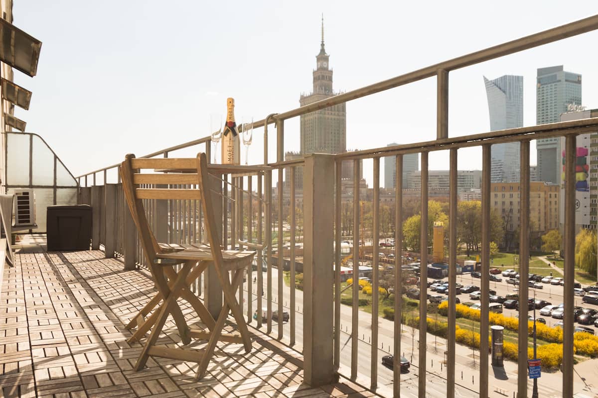 Un balcón con una silla de madera, barandilla metálica y vista a un paisaje urbano que incluye una torre destacada y edificios modernos.