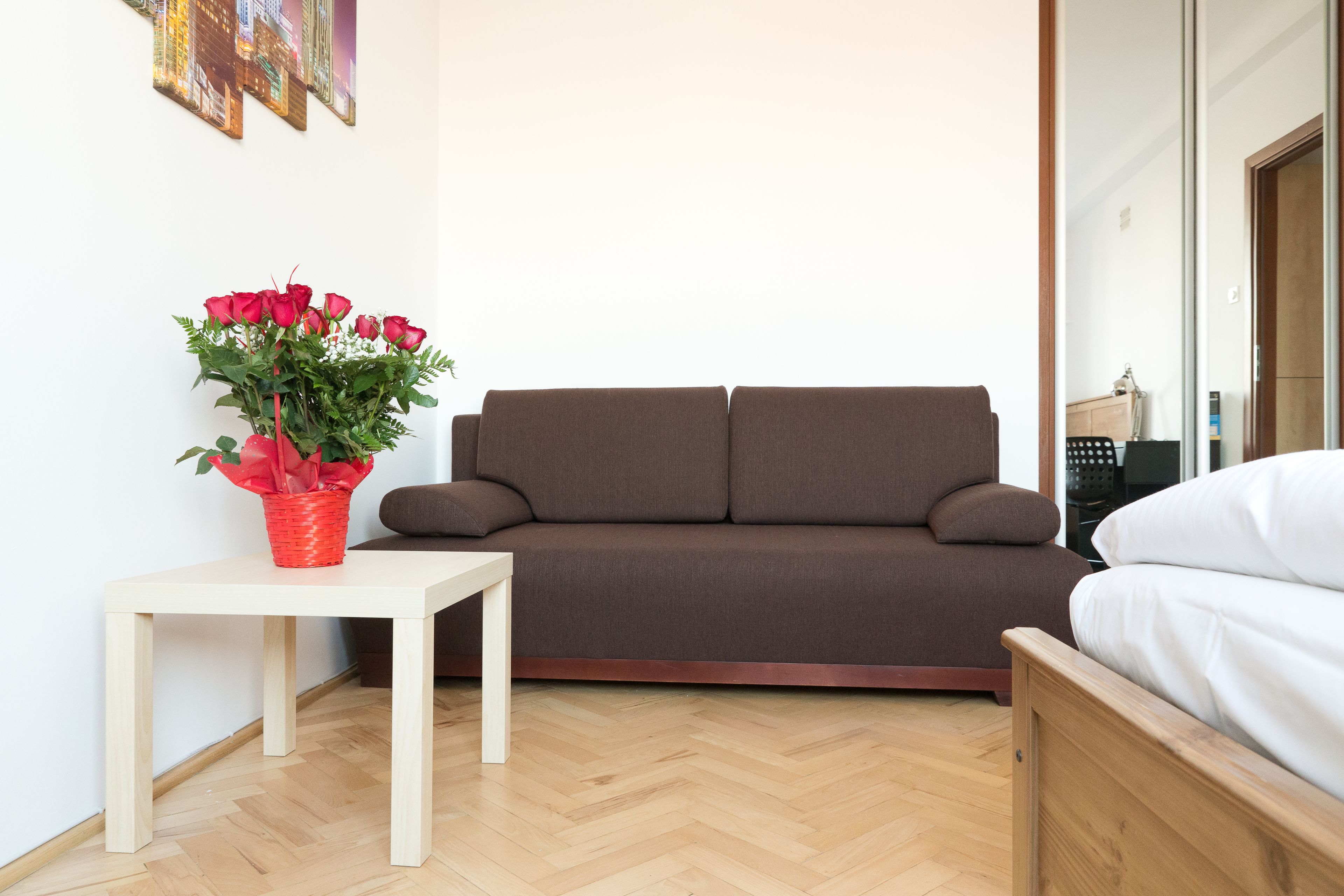 Una habitación moderna con un sofá marrón, una mesa blanca con un jarrón rojo de flores y una cama de madera.