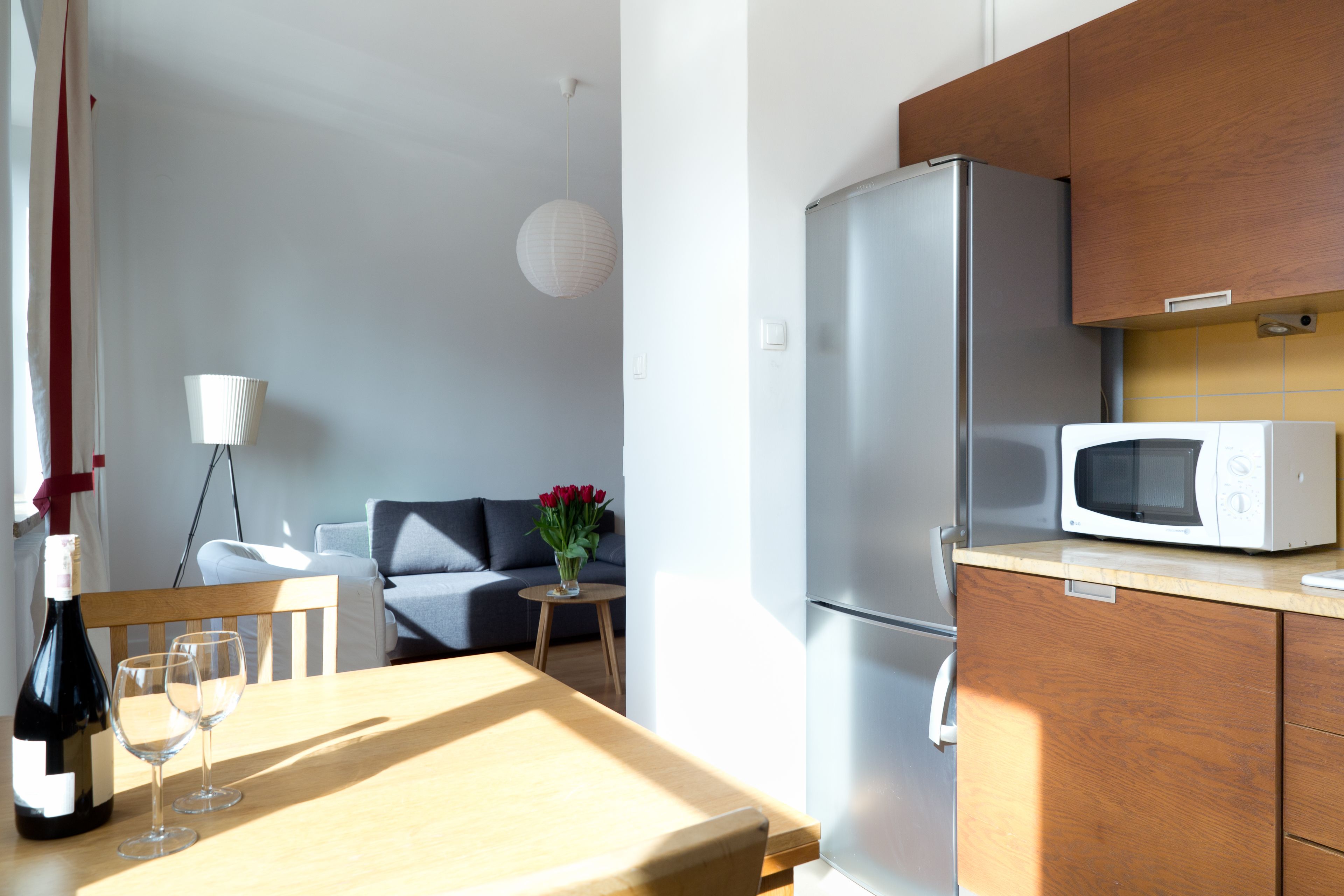 A modern kitchen with a refrigerator, microwave, wooden cabinets, and a dining table with wine glasses.