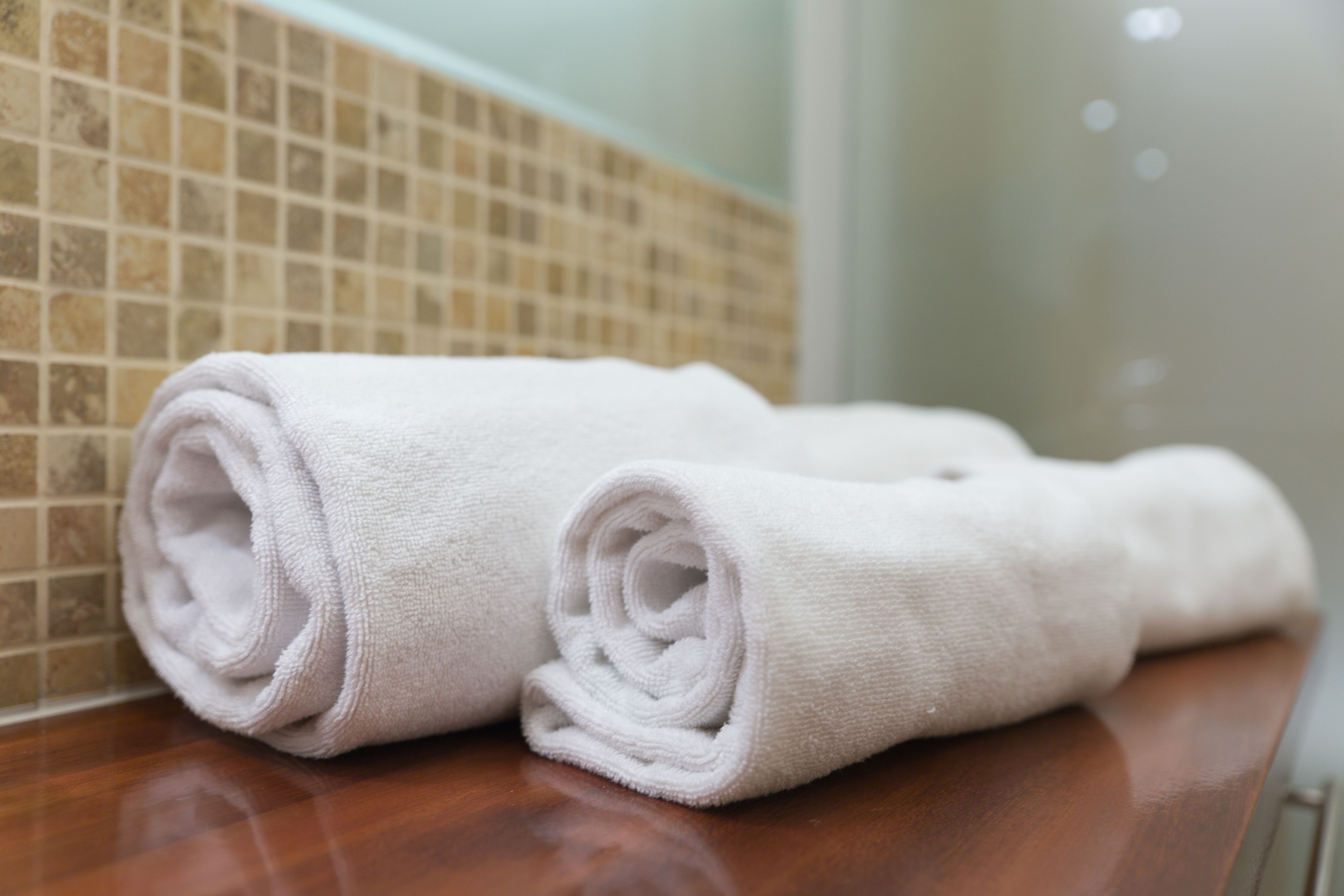 Two rolled white towels on a wooden surface in a bathroom with tiled walls.